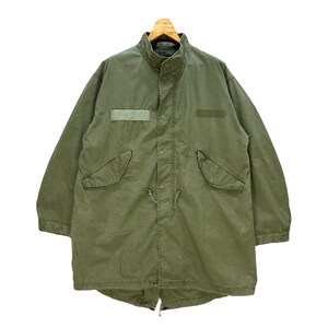 May include: Olive green military-style parka jacket with a high collar, two front pockets, and a drawstring waist. The jacket has a long length and is hanging on a wooden hanger. The jacket has two rectangular patches on the front.