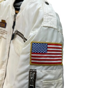 Pu&ograve; includere: Giacca bomber bianca con una toppa della bandiera americana sulla manica. La giacca ha una chiusura a zip, una piccola tasca con zip e una toppa con testo. La giacca &egrave; realizzata in materiale lucido.