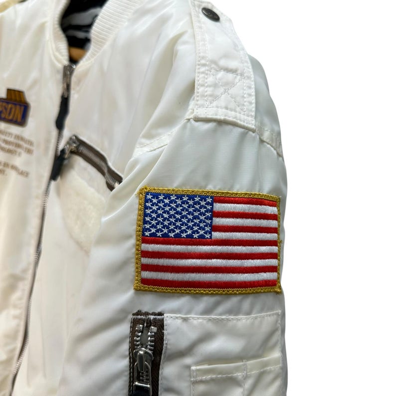 Pu&ograve; includere: Giacca bomber bianca con una toppa della bandiera americana sulla manica. La giacca ha una chiusura a zip, una piccola tasca con zip e una toppa con testo. La giacca &egrave; realizzata in materiale lucido.