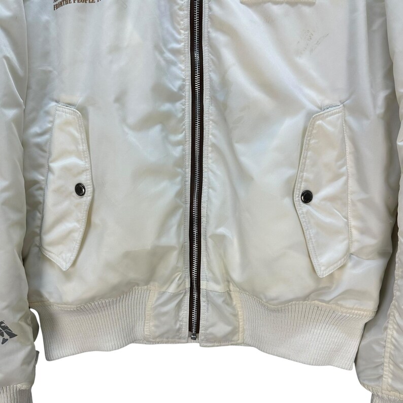 Pu&ograve; includere: Giacca bomber color crema con chiusura a zip completa e due tasche frontali. La giacca presenta orlo e polsini a coste e il testo "FROM THE PEOPLE" &egrave; visibile sulla parte superiore.