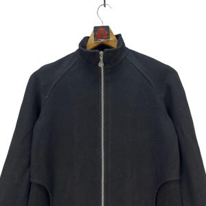 Peut inclure: Veste noire zipp&eacute;e avec un col montant. La veste a une fermeture &eacute;clair sur toute la longueur et un petit logo sur le curseur. La veste est suspendue &agrave; un cintre en bois.