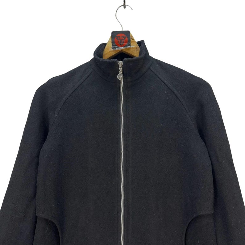Peut inclure: Veste noire zipp&eacute;e avec un col montant. La veste a une fermeture &eacute;clair sur toute la longueur et un petit logo sur le curseur. La veste est suspendue &agrave; un cintre en bois.