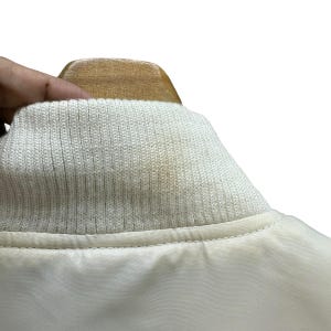 Pu&ograve; includere: Primo piano del colletto di un bomber color crema. Il colletto &egrave; a coste e il corpo della giacca &egrave; in tessuto liscio e chiaro. La giacca &egrave; appesa a un'appendiabiti di legno.