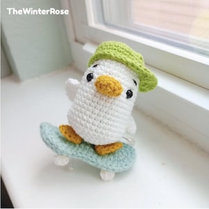 Puede incluir: Un pájaro blanco de crochet que lleva un sombrero verde y monta un monopatín azul. El pájaro tiene ojos amarillos y un pico amarillo.