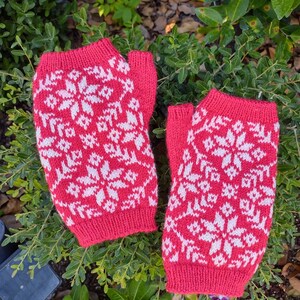 Puede incluir: Un par de guantes sin dedos de punto rojo con un patrón de copo de nieve blanco. Los guantes están hechos de un hilo suave y texturizado. Los guantes están diseñados para mantener las manos calientes. Se muestran sobre follaje verde.