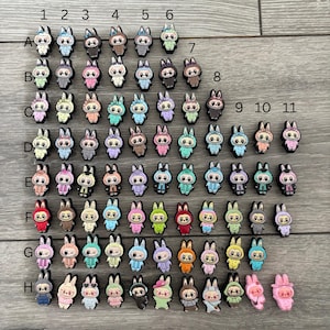 May include: A selection of colourful bunny-shaped charms. Each charm has a unique outfit and expression, with variations in colour and design. The charms are arranged in rows and columns, showcasing a variety of styles and colours.
