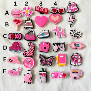 Könnte beinhalten: Ein Set mit 24 pinken und weißen Schuh-Anhängern mit verschiedenen Designs, darunter ein Herz, ein Schmetterling, eine Kamera, ein Blitz, eine Blume, eine Sonnenbrille, ein Koffer, eine Tasse Tee, ein Lollipop, ein Auto, eine Handtasche, ein Tintenfisch, ein Leopard, eine Schlafmaske, ein Milchpaket, ein Wal, ein Paar Hände, ein Ballontier, ein Paar Lippen, ein Macaron, ein Cowboyhut, ein 'XOXO'-Anhänger, ein 'BAE'-Anhänger, ein 'GRL PWR'-Anhänger und ein 'GIRLS'-Anhänger.