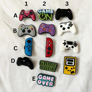 May include: A set of 10 colourful shoe charms featuring video game themes. The charms include a blue and red Nintendo Switch, a purple and green controller, a black and white controller, a black controller with the words "Game On", a green controller with the words "Game Over", a yellow Game Boy, and a black controller with the words "Game Over".
