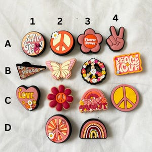 Może przedstawiać: Zestaw 12 kolorowych, inspirowanych pokojem charmów o różnych wzorach, w tym symbole pokoju, kwiaty, motyle, serca, tęcze i napisy "Stay Groovy", "Peace Love" i "Star Groovy".