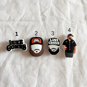 May include: Four different Luke Combs shoe charms. The first charm is a black and white logo with the text "LUKE COMBS". The second charm is a cartoon of Luke Combs wearing a black and white baseball cap with the text "LUKE COMBS" on it. The third charm is a cartoon of Luke Combs wearing a black and white baseball cap with the text "LUKE COMBS" on it. The fourth charm is a cartoon of Luke Combs wearing a black shirt and a black baseball cap. He is holding a red cup.