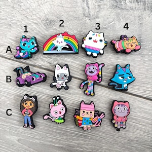 May include: A selection of colourful cartoon cat charms, including a blue cat with a bow, a cat on a rainbow, and a white cat with a sweater. Other charms include a purple race car and a girl.