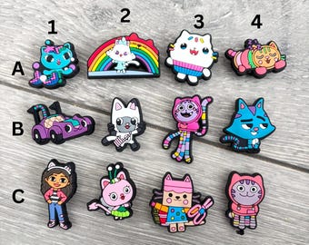 May include: A collection of colorful cartoon cat charms, including a blue cat with a bow, a cat on a rainbow, and a white cat with a sweater. Other charms include a purple race car and a girl.