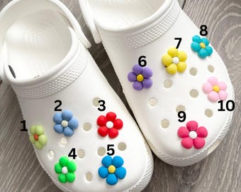 May include: White clogs adorned with colorful flower charms. The clogs feature various flower designs in shades of green, blue, red, yellow, purple, and pink. The flowers are attached to the top of the clogs, adding a decorative touch.