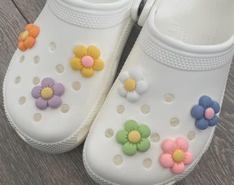 May include: A pair of white clogs with colorful flower embellishments. The flowers are in shades of pink, yellow, green, blue, and purple.