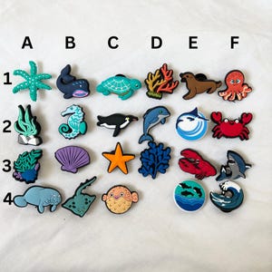 May include: A set of 24 colorful sea-themed charms, including a starfish, whale, turtle, coral, octopus, seahorse, penguin, dolphin, marlin, crab, seashell, star, seaweed, lobster, shark, pufferfish, stingray, and ocean waves. The charms are arranged in rows labeled A through F and 1 through 4.