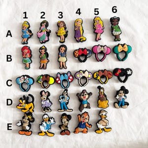 Peut inclure: Un ensemble de 25 breloques de chaussures de personnages Disney colorés, comprenant des princesses, Mickey Mouse et Minnie Mouse. Les breloques sont disposées en rangées étiquetées de A à E et de 1 à 6.