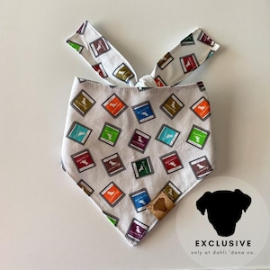 May include: White bandana with a colorful pattern of small squares featuring a dog silhouette. The bandana has a brown leather patch with the text "EXCLUSIVE only at dahli 'dana co."