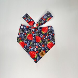 May include: A blue bandana with a white background featuring a pattern of red strawberries, blackberries, and blueberries. The bandana has a white border and a brown label.