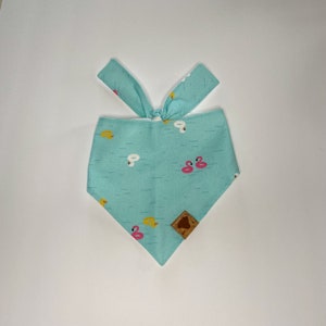 May include: A turquoise bandana with a pattern of pink flamingos and yellow rubber ducks on a blue wave background. The bandana has a brown leather patch with a logo on it.