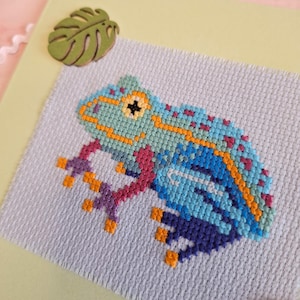 May include: A cross-stitch frog in shades of blue, teal, and purple, with orange accents. The frog is on a light gray fabric, set against a pale yellow background. A green leaf-shaped embellishment is in the upper left corner.
