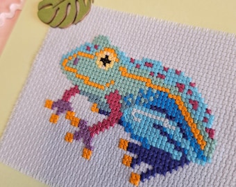 Könnte beinhalten: Ein Kreuzstichfrosch in Blau-, Türkis- und Lilatönen mit orangefarbenen Akzenten. Der Frosch befindet sich auf einem hellgrauen Stoff, vor einem hellgelben Hintergrund. Eine grüne, blattförmige Verzierung befindet sich in der oberen linken Ecke.