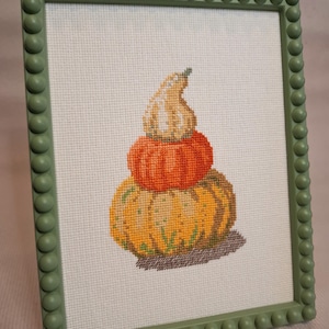 May include: Cross-stitch art of three stacked pumpkins in shades of orange, yellow, and tan, set against a white background. The artwork is framed in a green frame with a beaded edge, creating a fall-themed decorative piece.