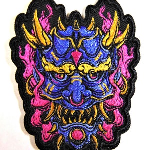 May include: A blue and gold embroidered patch of a Japanese demon mask with pink flames. The mask has a fierce expression with sharp teeth and horns.