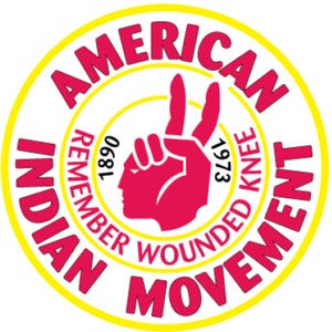 May include: A yellow circle with the words "American Indian Movement" in red around the outside. Inside the circle is a red hand making a peace sign with the words "Remember Wounded Knee 1973 1890" in black.