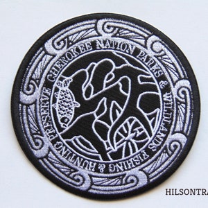 May include: A black and white embroidered patch with a circular design. The patch features the text "Cherokee Nation Parks & Wildlife" and a stylized image of a fish and a tree. The patch is surrounded by a decorative border.