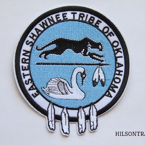 May include: A white and black embroidered patch with the text "Eastern Shawnee Tribe of Oklahoma" in a circle around a blue background. The patch features a black panther leaping over a white swan with a spear and feathers. The bottom of the patch has three white feathers.