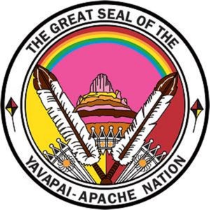 May include: The Great Seal of the Yavapai-Apache Nation features a rainbow arch over a pink mountain with a yellow and brown layered base. Two white feathers with black tips are crossed in front of the mountain. The seal is surrounded by a black circle with the text "The Great Seal of the Yavapai-Apache Nation" in white.