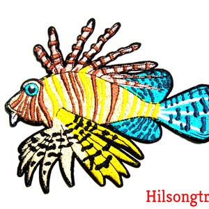 May include: An embroidered iron-on patch of a lionfish with yellow, brown, and blue stripes. The lionfish has a large, spiky fin and a small, pointed mouth.
