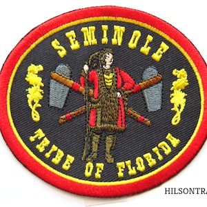 May include: A black and yellow embroidered patch with a red border. The patch features a Seminole Tribe of Florida logo with a Native American figure holding a tomahawk and a spear. The text "Seminole Tribe of Florida" is embroidered in yellow around the figure.