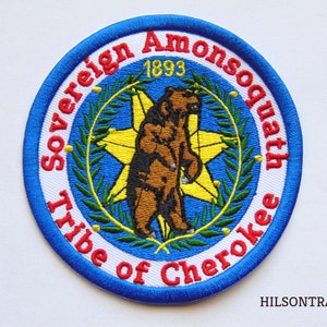 May include: A blue and white embroidered patch with the text "Sovereign Amonsquath Tribe of Cherokee 1893" around the edge. A brown bear stands in the center of the patch, surrounded by a yellow star and green leaves.
