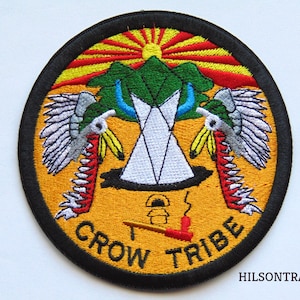 May include: A round, embroidered patch with a yellow background and black border. The patch features a stylized image of a teepee, a sun, and two birds with wings spread. The text "CROW TRIBE" is written in a black, curved font around the bottom of the patch.