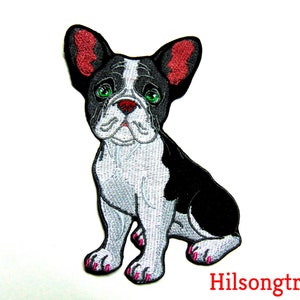 May include: Black and white embroidered patch of a French bulldog sitting with pink nail polish. The dog has green eyes and a red nose.