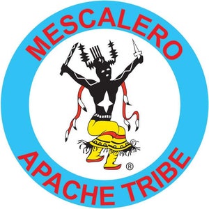 May include: A blue circle with the words "Mescalero Apache Tribe" in red around the edge. In the center is a black silhouette of a Native American dancer with a yellow skirt and red and white accents. The dancer is holding a knife in one hand and a stick in the other.