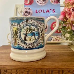 May include: A cream-colored ceramic mug with a blue and white design featuring penguins and the text "Warming Explorers on Polar Expeditions 1909." The mug has a curved handle and a decorative border. The mug is sitting on a wooden surface.