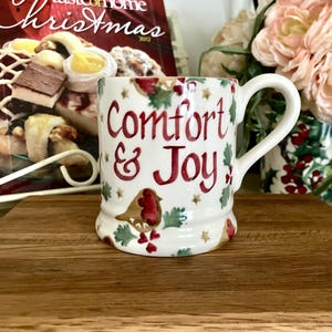 May include: White ceramic mug with the words "Comfort & Joy" in red script. The mug features a robin and holly design, with gold stars. A Christmas-themed magazine is in the background.