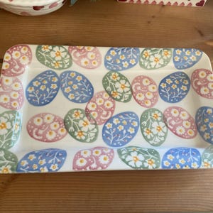 May include: Rectangular ceramic serving tray with a white base. The tray is decorated with a repeating pattern of pastel-colored Easter egg shapes, each adorned with a floral design. The eggs are in shades of blue, pink, and green.