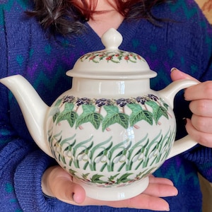 Może przedstawiać: Kremowy ceramiczny czajniczek z ręcznie malowanym wzorem botanicznym. Projekt zawiera rzędy zielonych liści bluszczu, ciemnych jagód, białych kwiatów przebiśniegów oraz małych czerwonych owoców ostrokrzewu zdobiących pokrywkę i korpus.