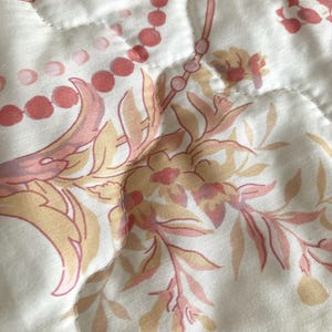 May include: Close-up of a quilted fabric with a floral pattern in shades of peach, pink, and beige. The design features stylized flowers, leaves, and beaded accents on a white background. The fabric has a soft, textured appearance.