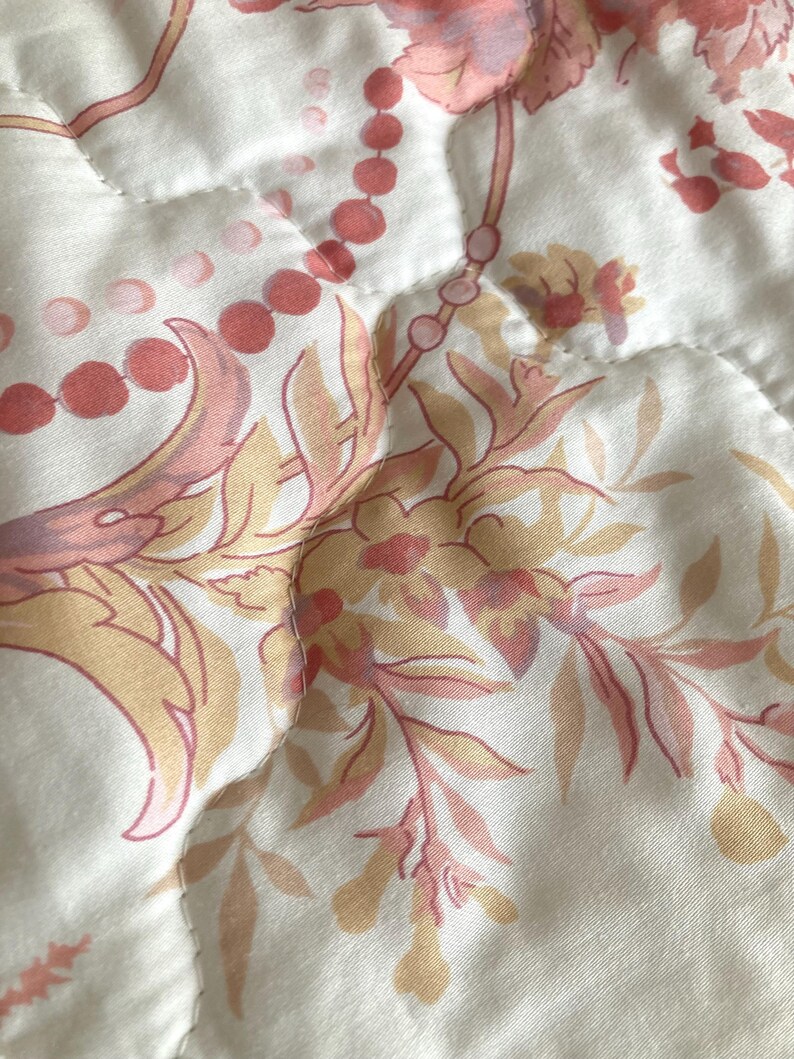 May include: Close-up of a quilted fabric with a floral pattern in shades of peach, pink, and beige. The design features stylized flowers, leaves, and beaded accents on a white background. The fabric has a soft, textured appearance.
