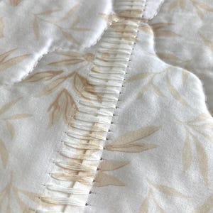 May include: Close-up of a quilted fabric with a floral pattern in shades of beige and white. The fabric features a central, textured seam with vertical stitching. The overall design suggests a bedding or home decor item.