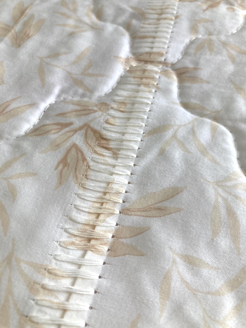 May include: Close-up of a quilted fabric with a floral pattern in shades of beige and white. The fabric features a central, textured seam with vertical stitching. The overall design suggests a bedding or home decor item.