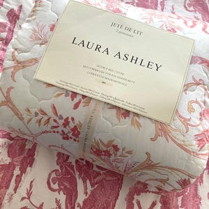 May include: A double bed cover by Laura Ashley, featuring a floral and pastoral design in shades of pink and cream. The cover is quilted and includes the text "JET&Eacute; DE LIT" and "DOUBLE BED COVER".