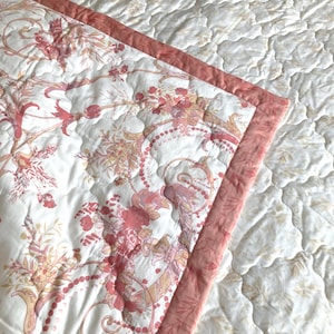May include: A quilted bedspread with a floral pattern in shades of pink, peach, and cream. The edge is trimmed with a solid pink border. The background is a white quilted fabric with a subtle leaf pattern.
