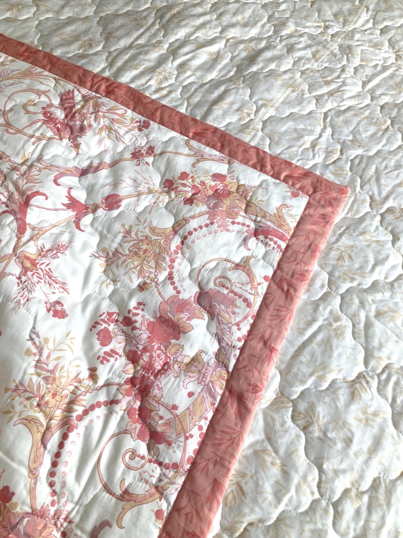 May include: A quilted bedspread with a floral pattern in shades of pink, peach, and cream. The edge is trimmed with a solid pink border. The background is a white quilted fabric with a subtle leaf pattern.