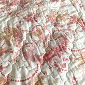May include: Close-up of a quilted fabric with a floral pattern in shades of pink, peach, and cream. The design features stylized flowers, leaves, and beaded accents, creating a vintage aesthetic. The fabric has a quilted texture.
