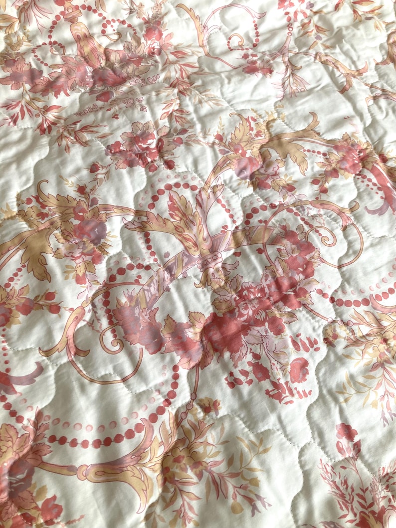 May include: Close-up of a quilted fabric with a floral pattern in shades of pink, peach, and cream. The design features stylized flowers, leaves, and beaded accents, creating a vintage aesthetic. The fabric has a quilted texture.